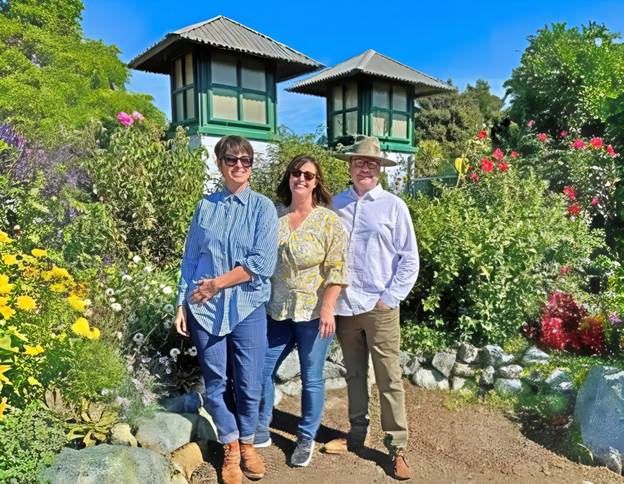 Three adults standing together in a vibrant garden filled with flowers and greenery, with two distinctive structures visible in the background.